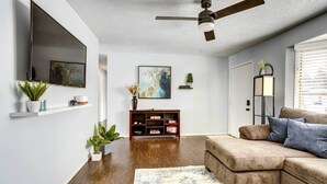 A living room with a brown sectional sofa, a ceiling fan, a mounted TV, a bookshelf, and a potted plant. - Cozy Sioux Falls Home - 7 Mi to Downtown! (Sioux Falls)