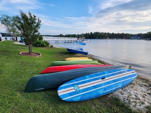 A lakeside view with a row of colorful kayaks lined up on the grass, a dock in the background, and a clear sky.
