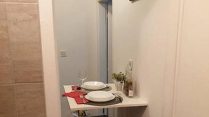 A small, narrow room with a tiled floor, a white wall, and a door leading to another room. There is a small table with a chair, a framed picture on the wall, and a red cloth on the table. - Iva - Modern Downtown - A2 Navada (Split)