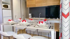Una cocina moderna con armarios de madera, un microondas y una cafetera. Hay un comedor con sillas blancas y una mesa preparada para cuatro.