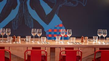 Un espace bar avec une longue table en bois, dressée avec des verres et des couverts, des chaises rouges et un luminaire suspendu rouge.