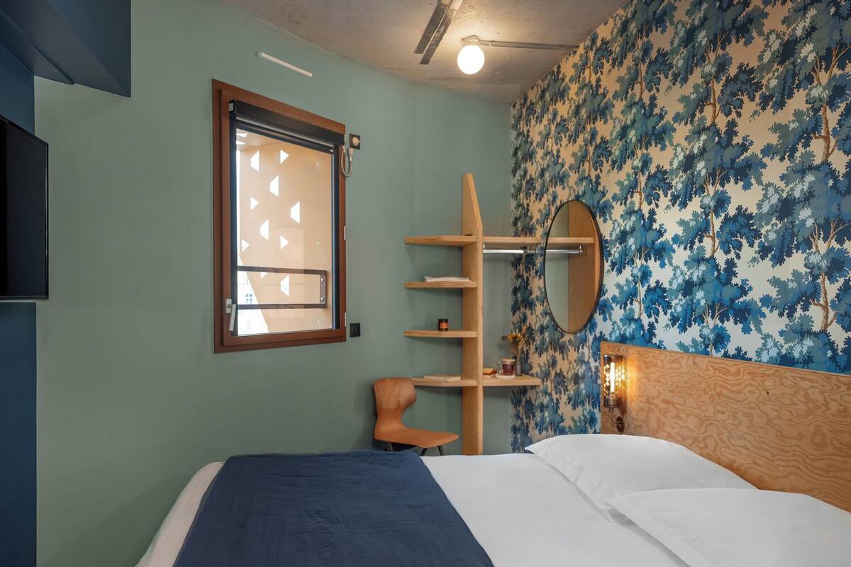 A bedroom with a bed, a window with blinds, a wooden shelf, a mirror, and a wall with a tree pattern.