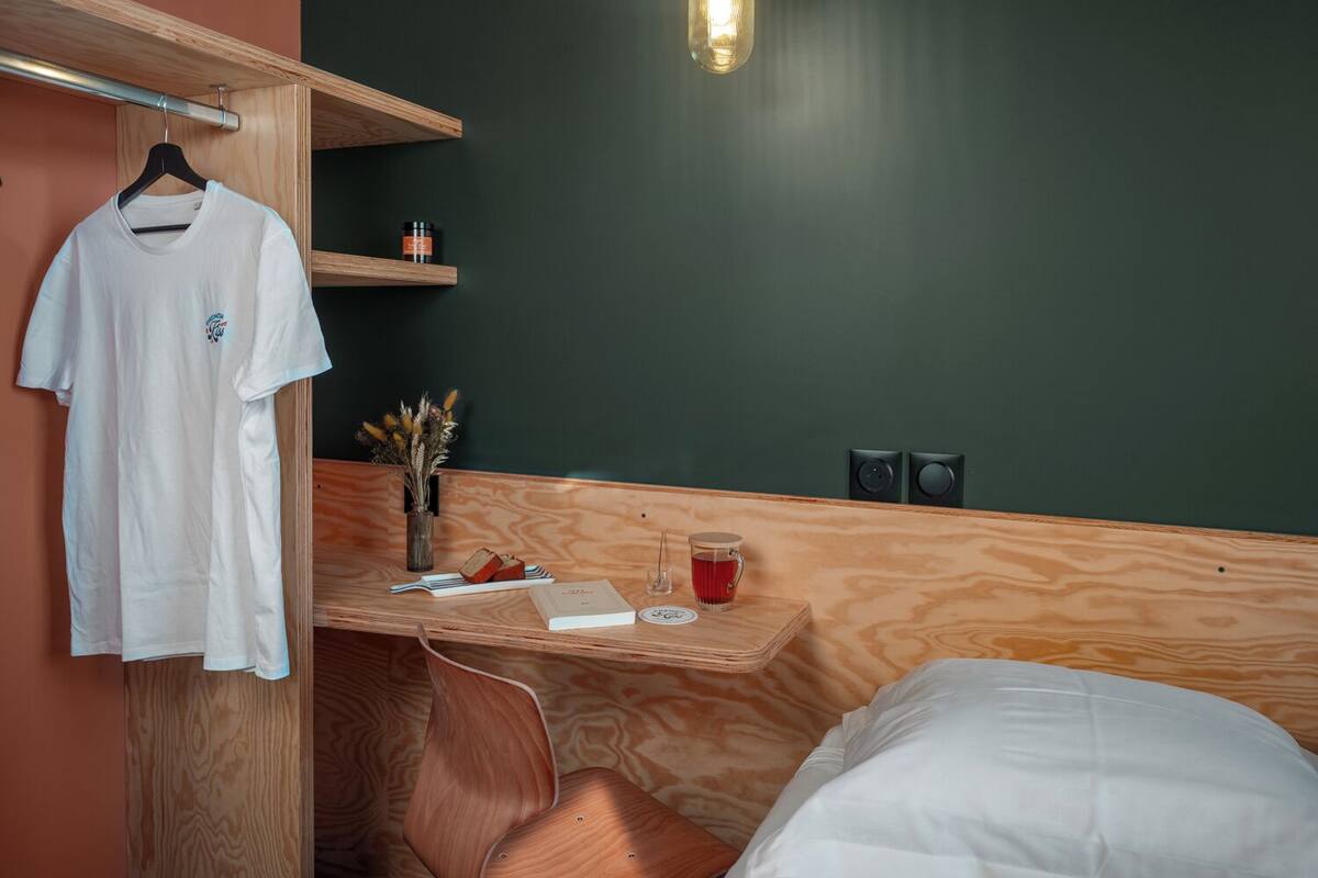 A room with a bed, a wooden desk, a hanging light fixture, a white t-shirt on a hanger, a vase with dried flowers, and two speakers on the wall.