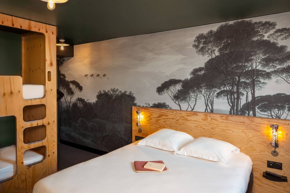 A bedroom with a bed, a wooden headboard, a mural of trees, and two wall-mounted lights.