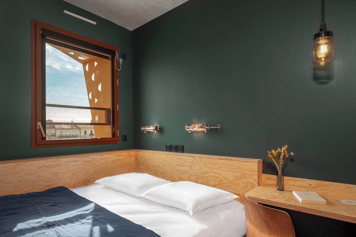 A hotel room with a bed, a desk with a chair, a window with a view, and wall-mounted lighting.