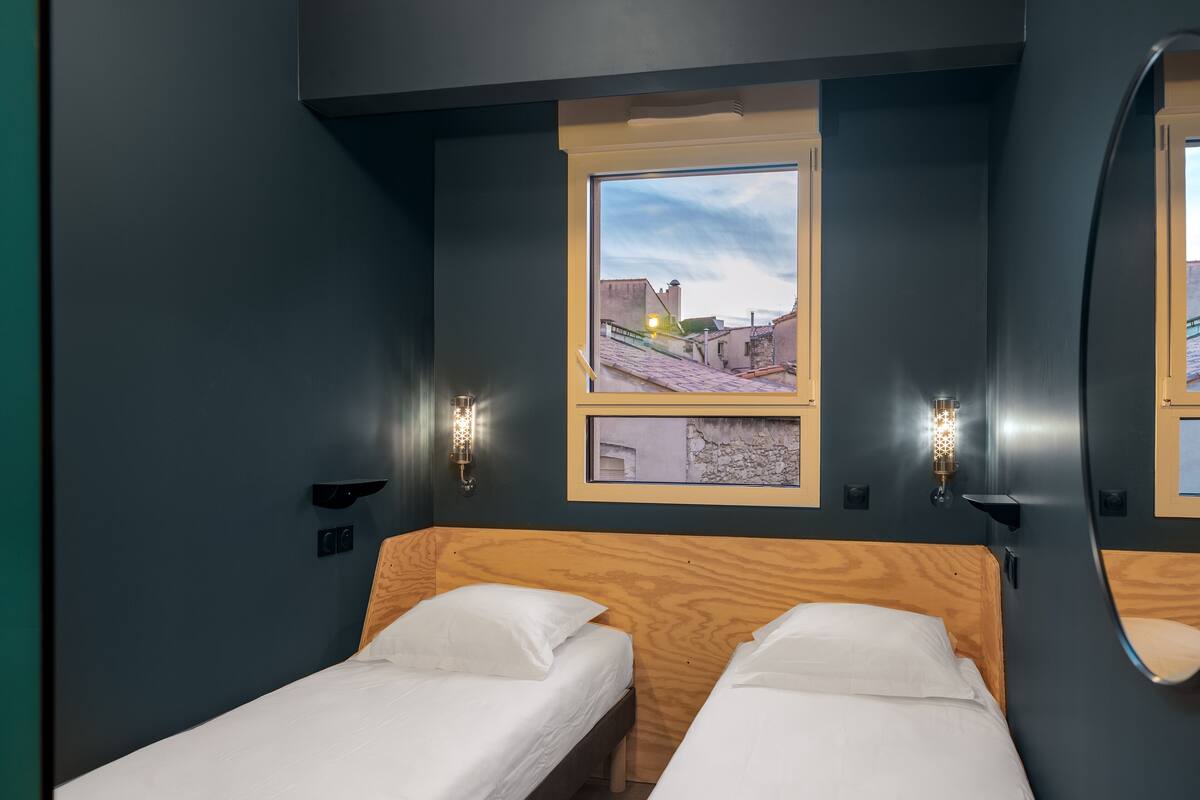 A small room with two single beds, a window with a view of buildings, and wall-mounted sconces.