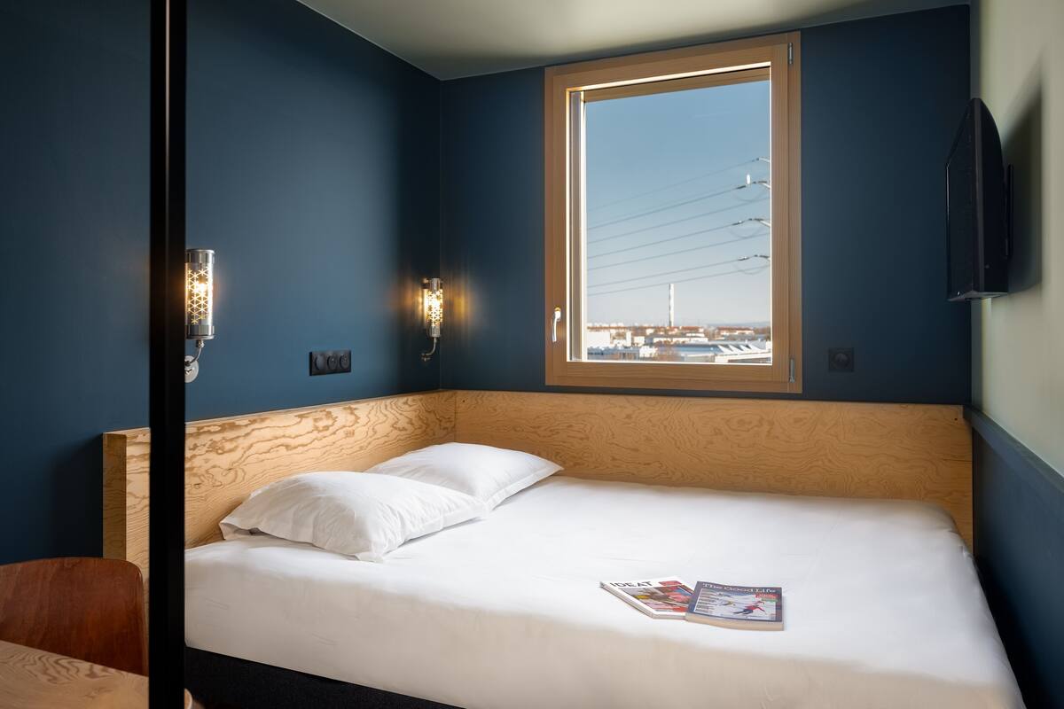 A hotel room with a bed, a window with a view of a cityscape, a wall-mounted TV, and two wall sconces.
