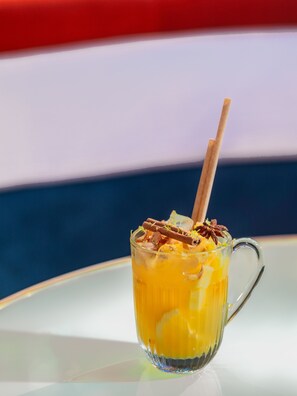 A glass mug with a straw, containing a yellow beverage and garnished with cinnamon sticks and star anise, placed on a white table with a blue and red striped background.