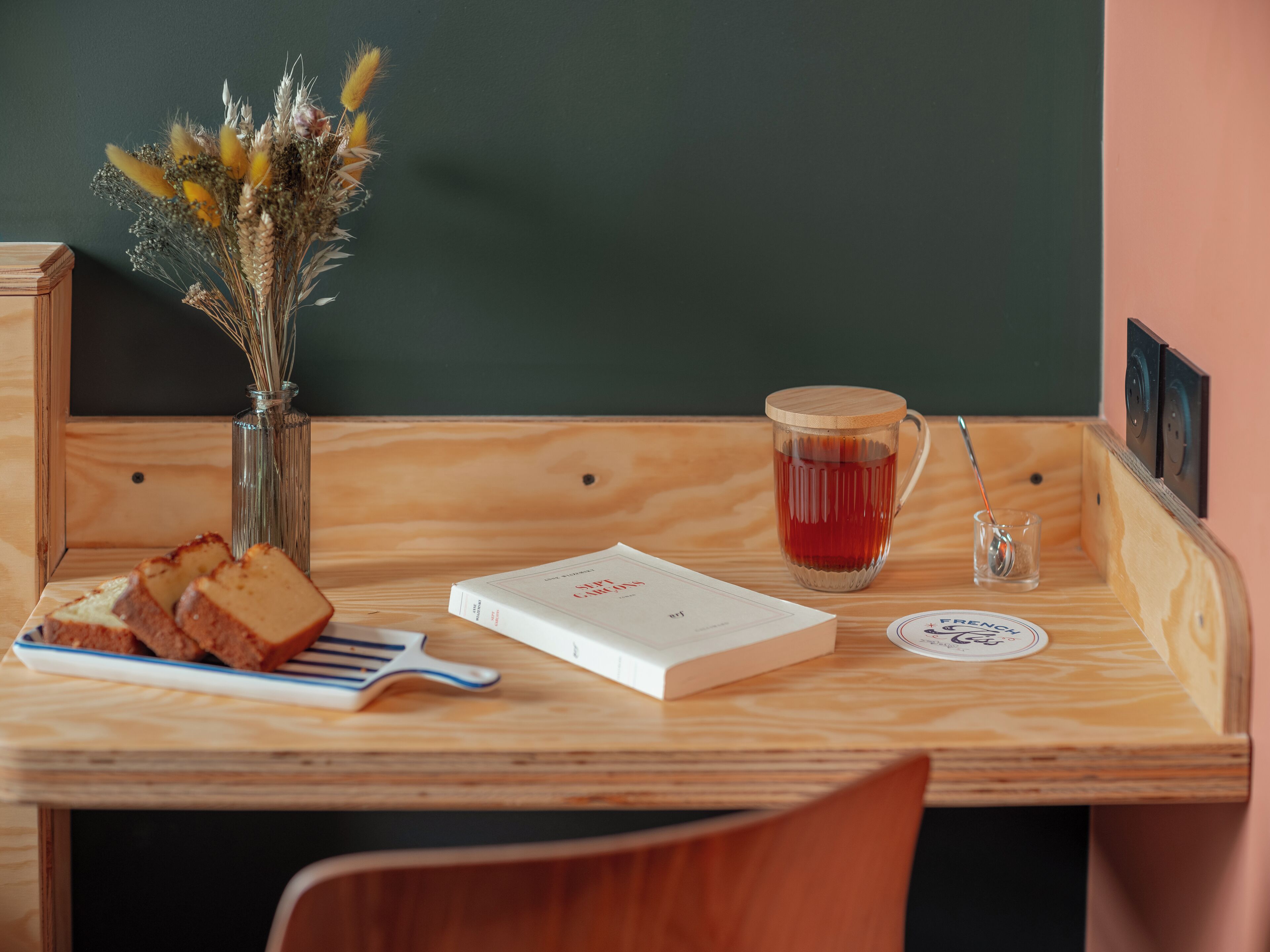 Ein Holztisch mit einem Buch, einer Tasse, einem Löffel und einer Vase mit getrockneten Blumen.