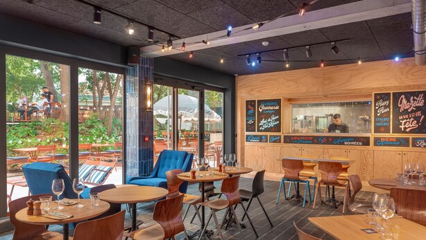 An indoor dining area with tables and chairs, large windows, and a wooden wall with menu boards. - Eklo Hotels Montpellier City center Saint-Roch train station (Montpellier)