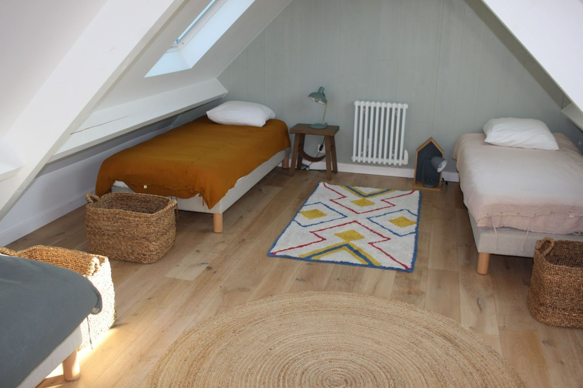 A small, cozy attic bedroom with a sloped ceiling, a bed with a brown blanket, a bedside table with a lamp, and a radiator.