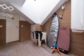 A small bathroom with a skylight, a toilet, a sink, and a surfboard leaning against the wall. - Dude Beach Hostel (Figueira da Foz)