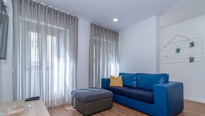 Uma sala de estar moderna com um sofá azul, um banquinho, uma mesa de centro de madeira e cortinas leves.