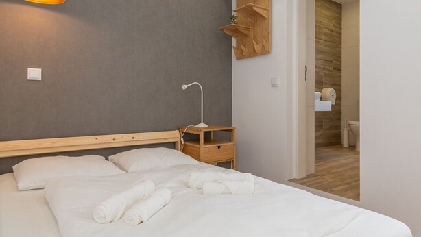 Cama bem arrumada com lençóis brancos e toalhas dobradas, mesa de cabeceira com abajur e cabeceira de madeira.
