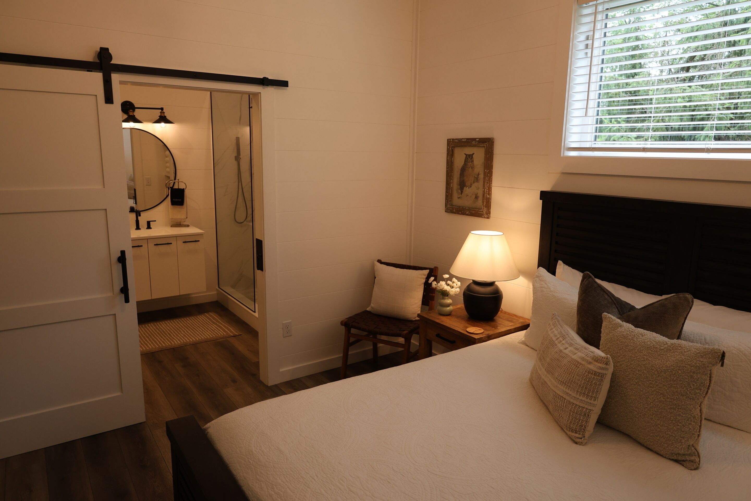 A bedroom with a bed, bedside table with a lamp, a chair, a window with blinds, and a door leading to a bathroom.