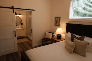 Un dormitorio con cama, mesita de noche con lámpara, una silla, ventana con persianas y puerta que da a un baño.