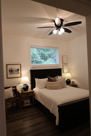 Quarto com cama, mesas de cabeceira, ventoinha no teto, janela e quadro na parede.