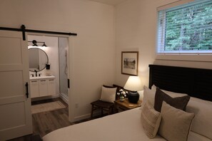 Un dormitorio con cama, mesita de noche, lámpara, ventana y puerta que da a un baño.