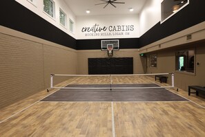 An indoor sports court with a basketball hoop, wooden flooring, and a ceiling fan.