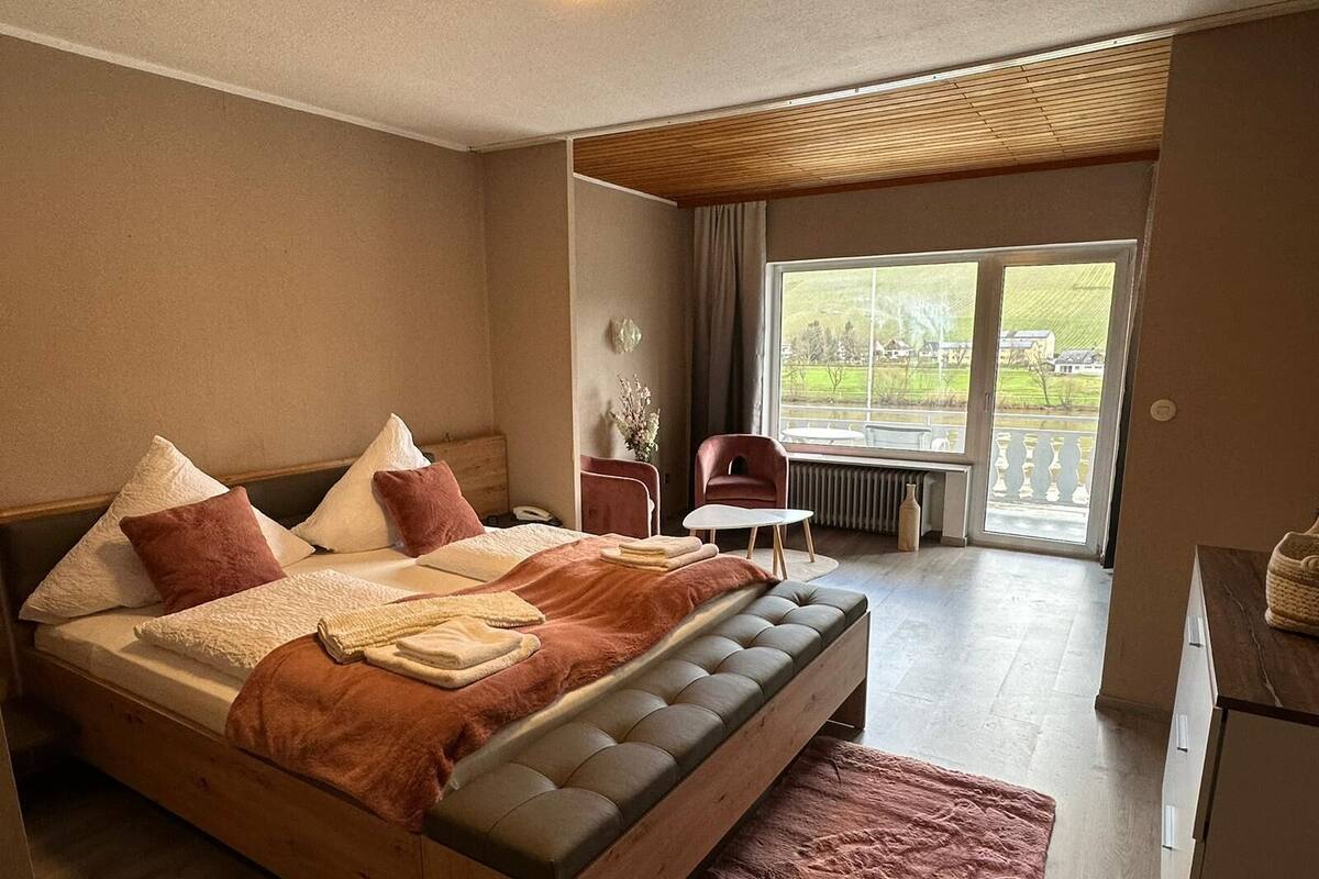 A spacious bedroom with a large bed, a small table, a chair, and a view of the outdoors through a large window.