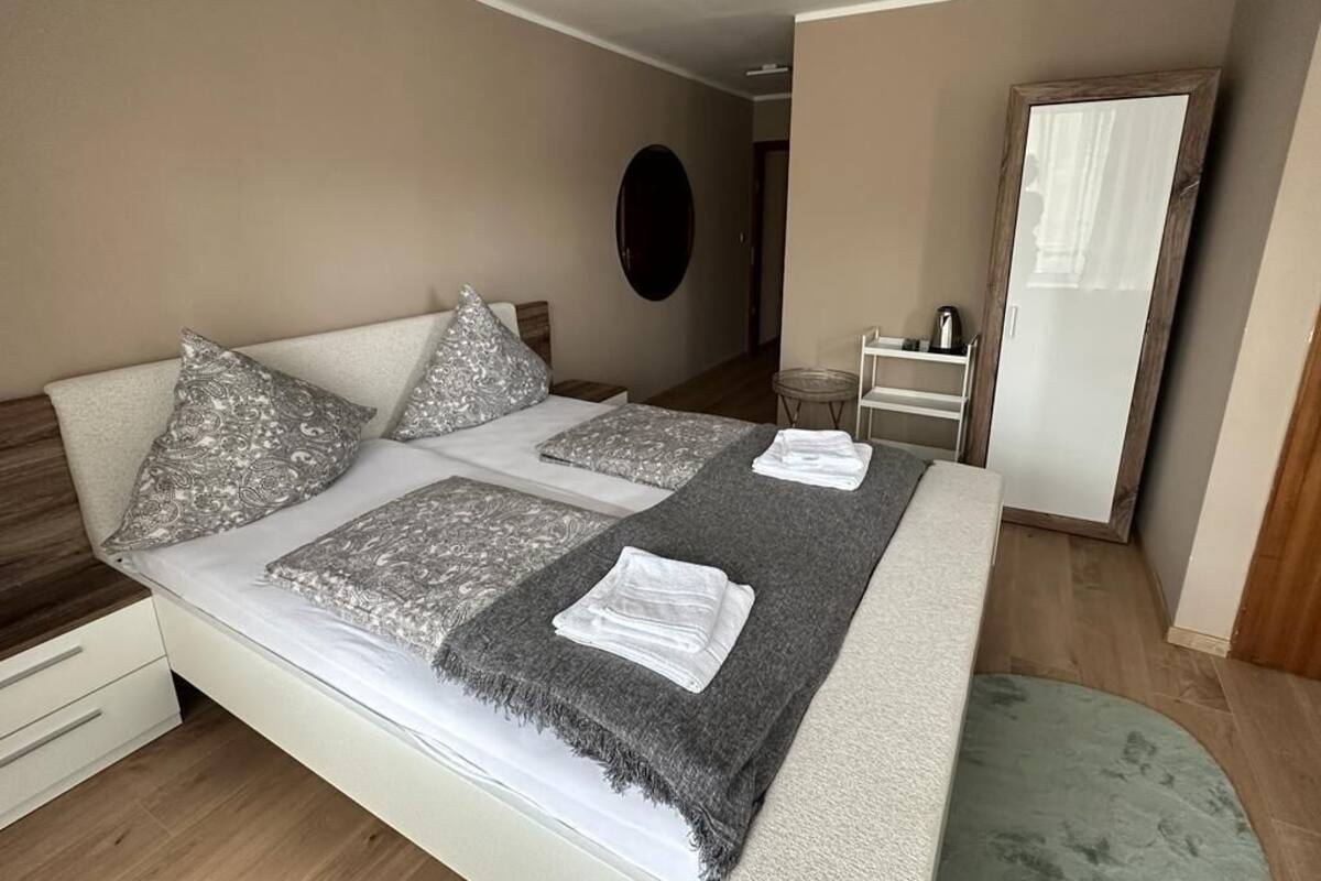 A neatly made bed with patterned pillows and a gray throw blanket, a small table with a kettle, and a mirror on the wall.