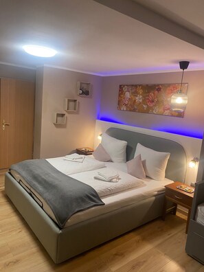 A neatly made bed with pillows and a blanket, a bedside table with a lamp, a wall-mounted shelf with a bicycle, and a painting above the bed.