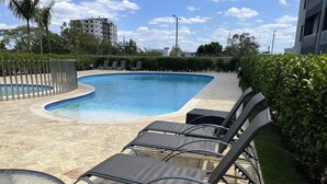 A swimming pool with lounge chairs and a table, surrounded by a hedge and a building in the background. - 10Min. Del Aeropuerto apt.picina aire wifi 24H seguridad patio de juegos parqueo (Santiago De Los Caballeros)