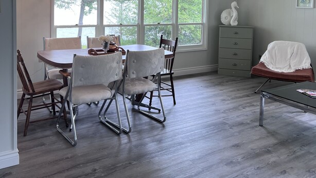A dining area with a round table and four chairs, large windows with a view of trees, a gray dresser, and a bench with a white cover. - Private getaway on Lower Buckhorn Lake (Trent Lakes)