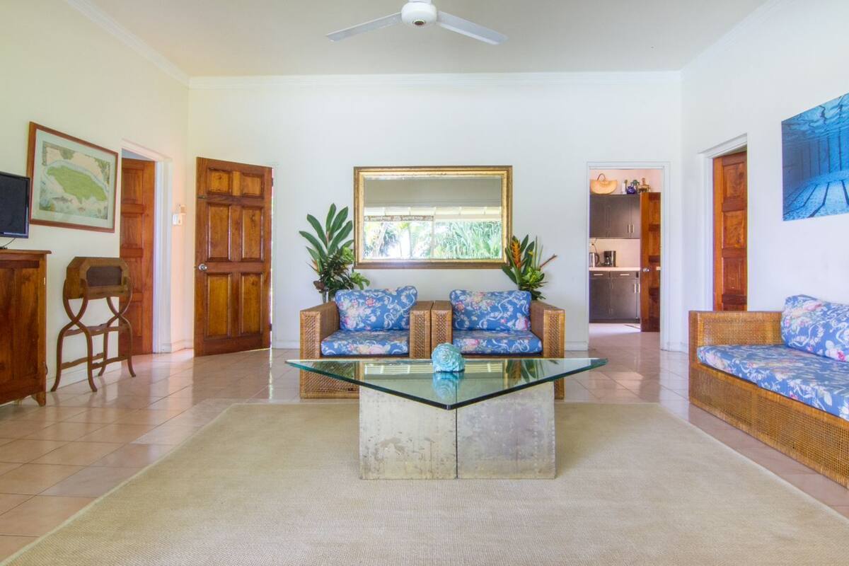 Un salón amplio con una mesa de centro de vidrio, dos sofás con tapicería floral, un armario de madera y una cocina visible a través de un umbral abierto.