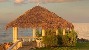A thatched-roof pavilion with a stone pathway leading to it, surrounded by greenery and overlooking a body of water.