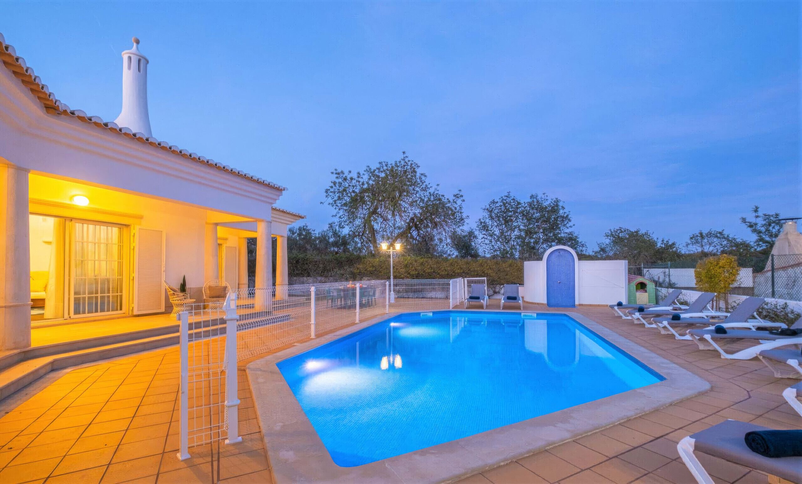 A swimming pool with a tiled deck, lounge chairs, and a white building with a chimney.