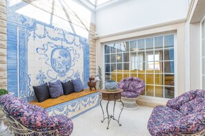 A room with a blue and white tiled wall, a wooden bench with cushions, a wicker chair, a metal table with a vase, and a large window. - Family retreat w/ heatable pool & BBQ (Albufeira)