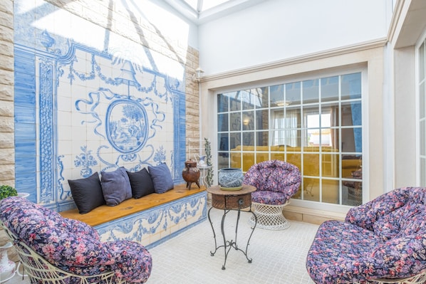 A room with a blue and white tiled wall, a wooden bench with cushions, a wicker chair, a metal table with a vase, and a large window.