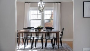 A dining area with a wooden table and chairs, a chandelier, and a view of a brick building through large windows. - Dundee's 3BR Upscale Retreat, Near Downtown! (Omaha)