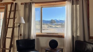 A room with a wooden ceiling, a window with curtains, two black chairs, a small table with a speaker, and a wooden ladder against the wall. - Cantina Stagecoach Stop (Grants)