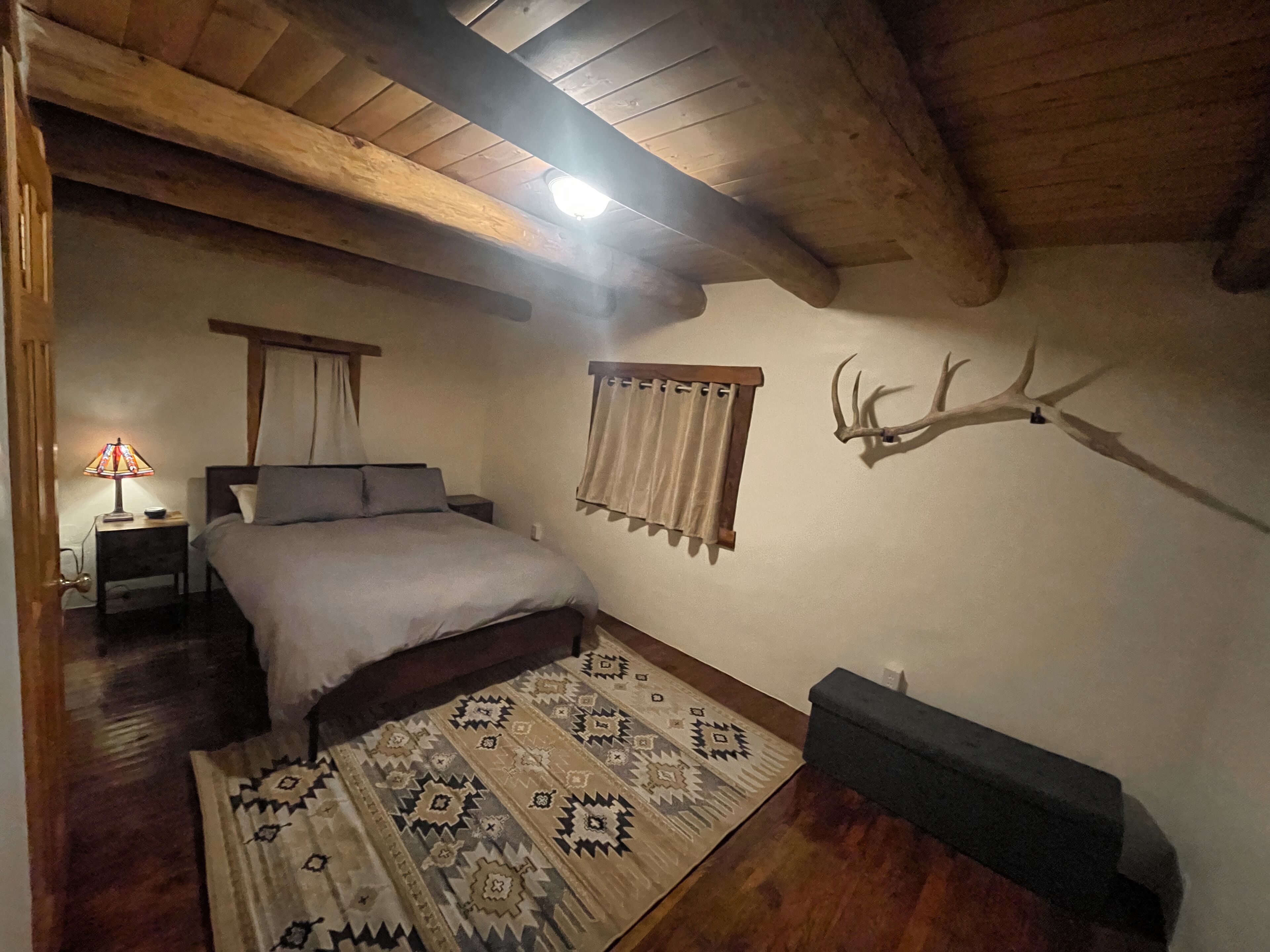 Un dormitorio con cama, un tapete estampado, una mesita de noche con lámpara, techo de madera y un cornamenta de ciervo montado en la pared.