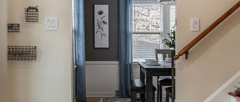 A hallway with a dining area, a staircase, and a window with curtains.