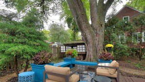 Een terras met tuinmeubilair, een grote boom en een huis op de achtergrond.