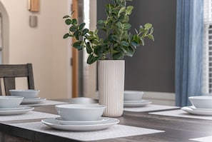 A dining table with white plates and bowls, a tall white vase with green foliage, and a dark wooden chair.