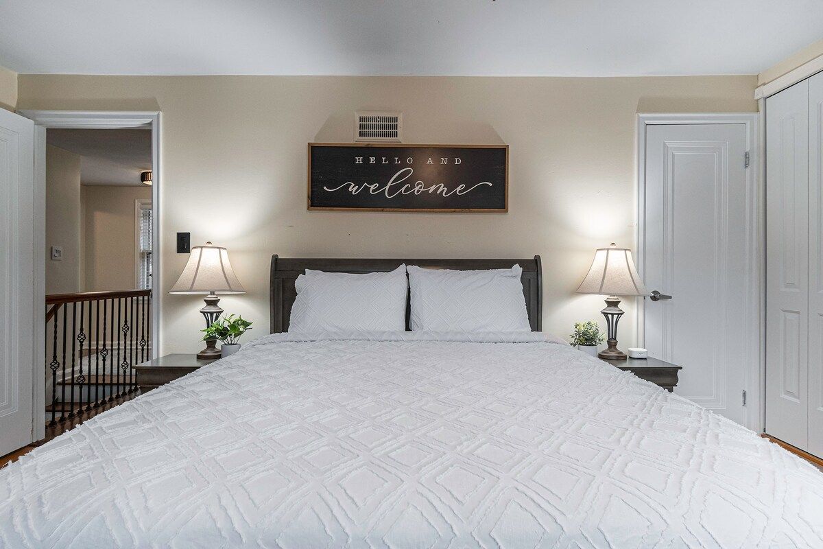 Une chambre avec un grand lit, deux lampes de chevet, une pancarte sur laquelle on peut lire « Bonjour et bienvenue », et une porte sur le côté droit.