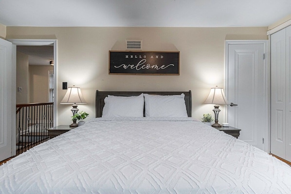 A bedroom with a large bed, two bedside lamps, a 'Hello and welcome' sign, and a door on the right.