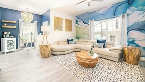 A living room with a curved sofa, a wooden coffee table, a ceiling fan, and a large window with curtains. - 5 BR Villa+Pool&Lake views+Near Disney+WaterPark (Kissimmee)