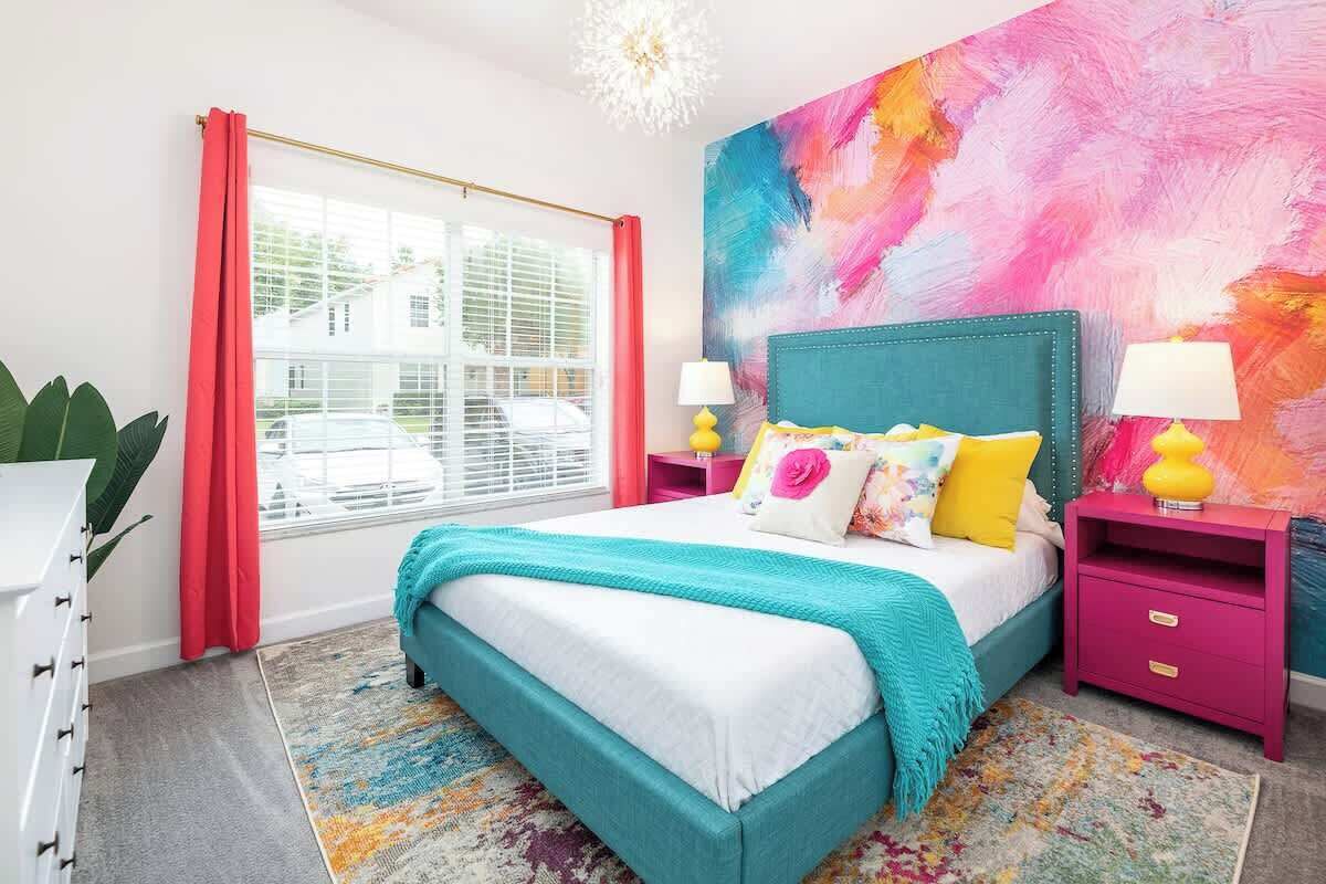 A bedroom with a bed, two bedside tables, a window with curtains, and a colorful abstract wall.