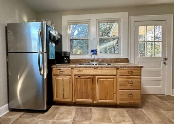 Compact kitchen with a refrigerator, sink, and coffee maker, featuring two windows and a tiled floor.