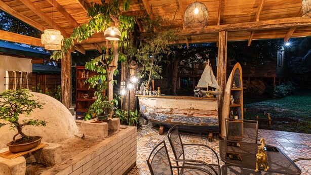 An outdoor patio with a brick wall, a wooden pergola, hanging wicker lamps, and a small fountain.
