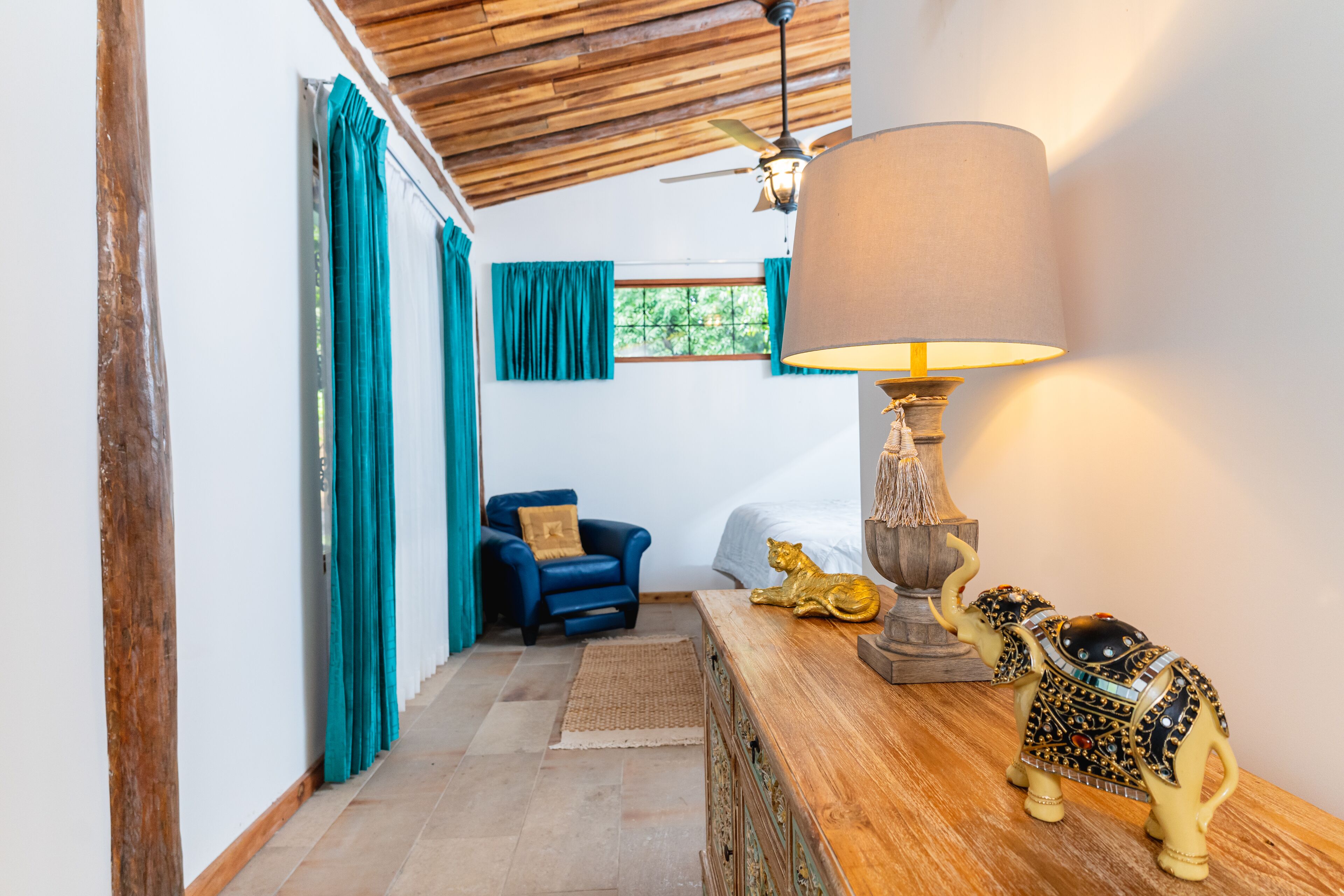 Ein Zimmer mit Holzbalkendecke, Deckenventilator, Bett, blauem Sessel, Lampe und einer dekorativen Elefantenskulptur.