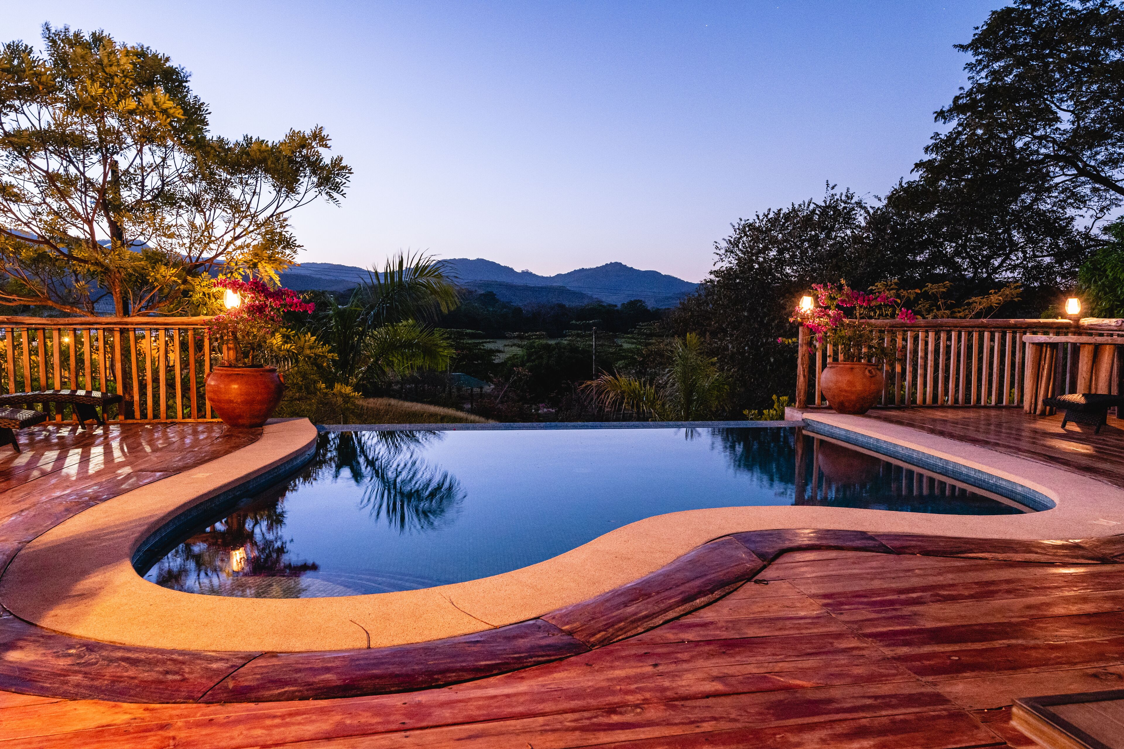 Une piscine dotée d’une terrasse en bois, entourée de plantes en pot et d’arbres, et offrant une vue sur les montagnes en arrière-plan.
