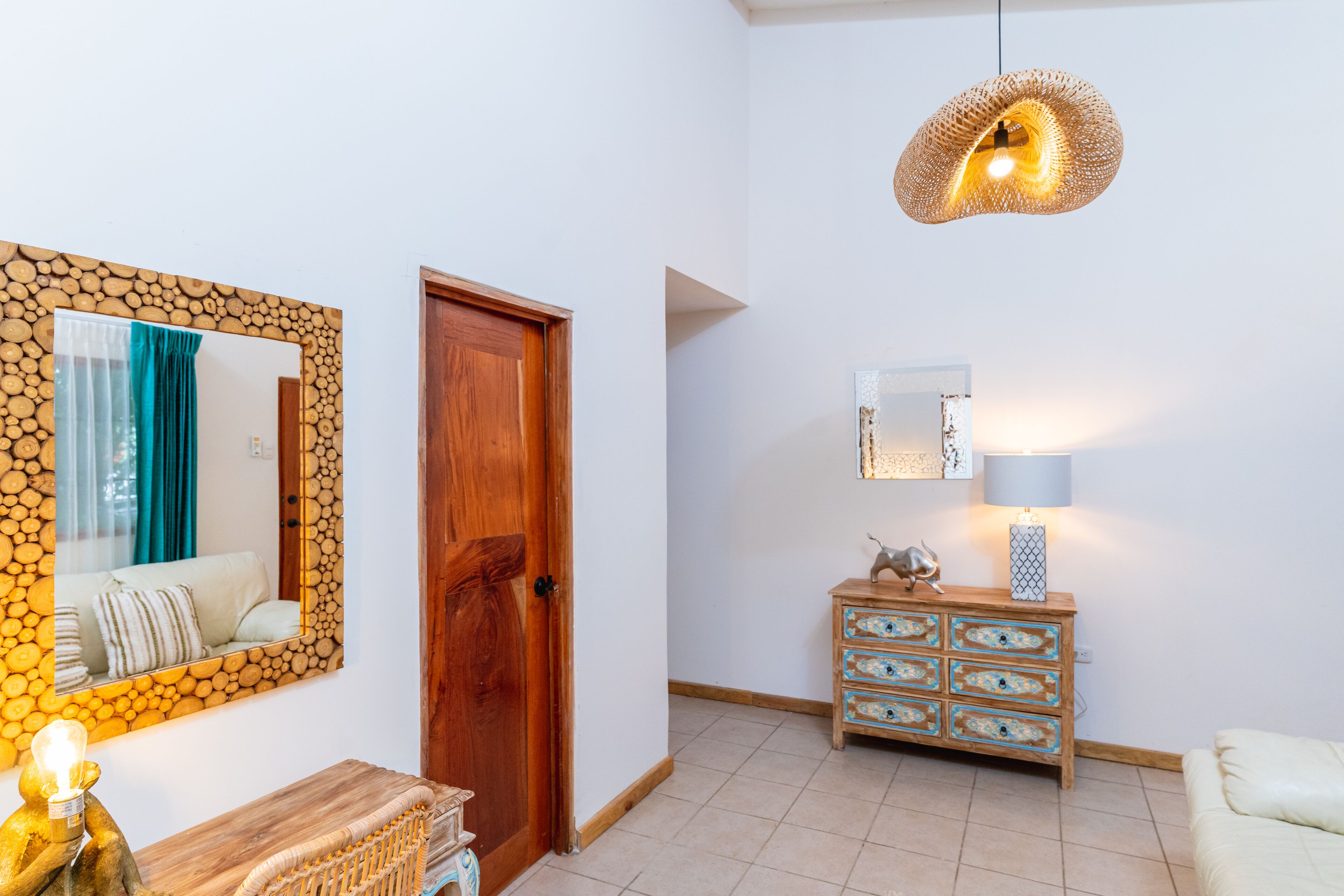 Ein Zimmer mit Holztür, einem Spiegel mit einem Rahmen im Blockbohlen-Stil, einer Holzkommode mit Schubladen, einer Pendelleuchte und einer Lampe auf der Kommode.
