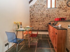 A small, stone-walled kitchen with a dining table, chairs, and a microwave on the counter. - The Threshing Barn (Beaworthy)