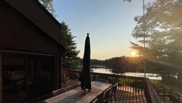Eine Holzterrasse mit Tisch und Stühlen, mit Blick auf einen See und einem Sonnenuntergang im Hintergrund.